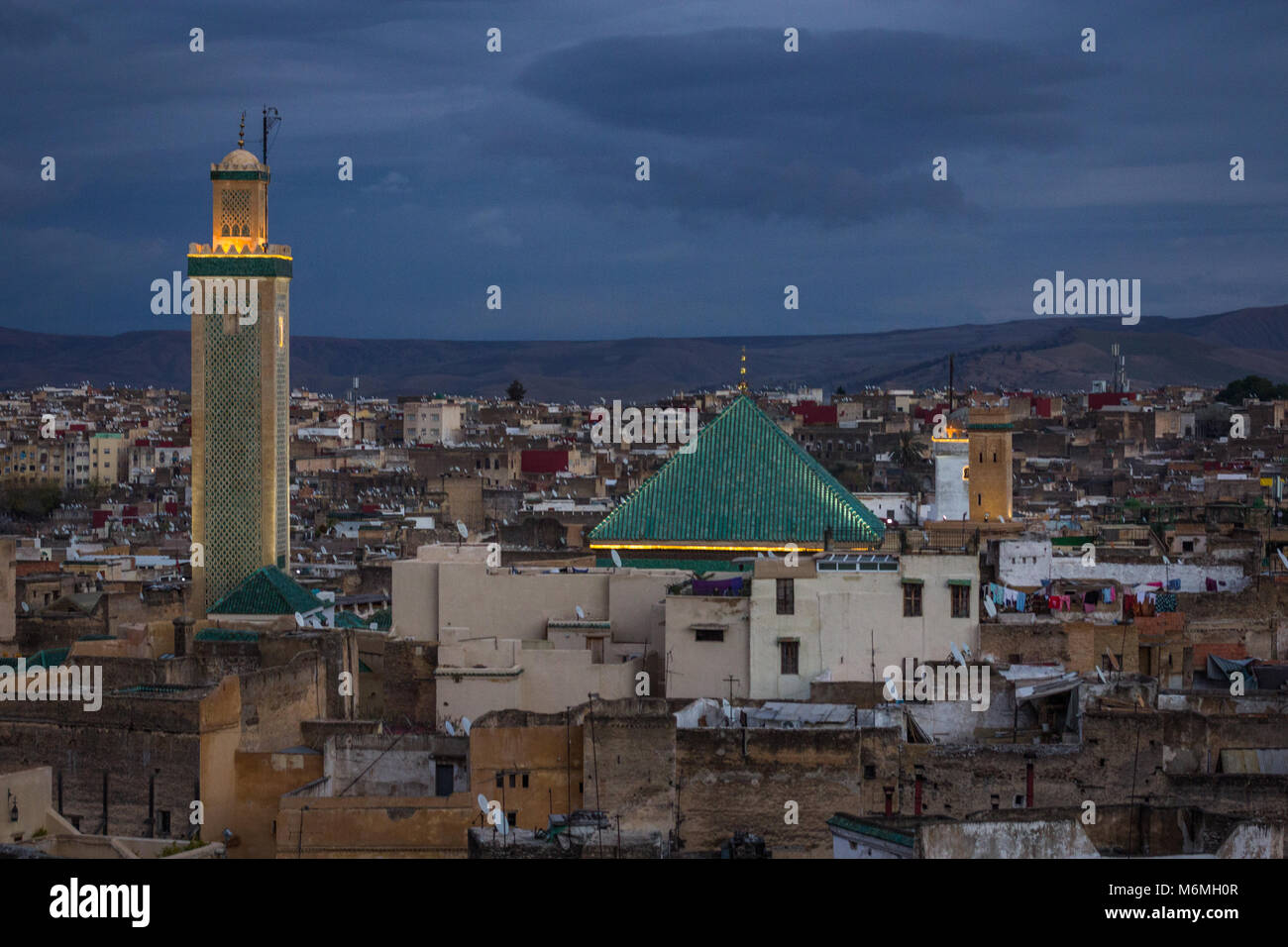 The lit minaret and rooftop of Karaouiyine Mosque at dusk, against the rooftops of Fes and the mountains in the background Stock Photo