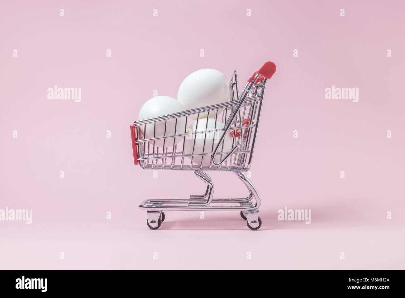 Shopping trolley model full of white eggs against pastel pink rose background minimal concept. Space for copy. Stock Photo