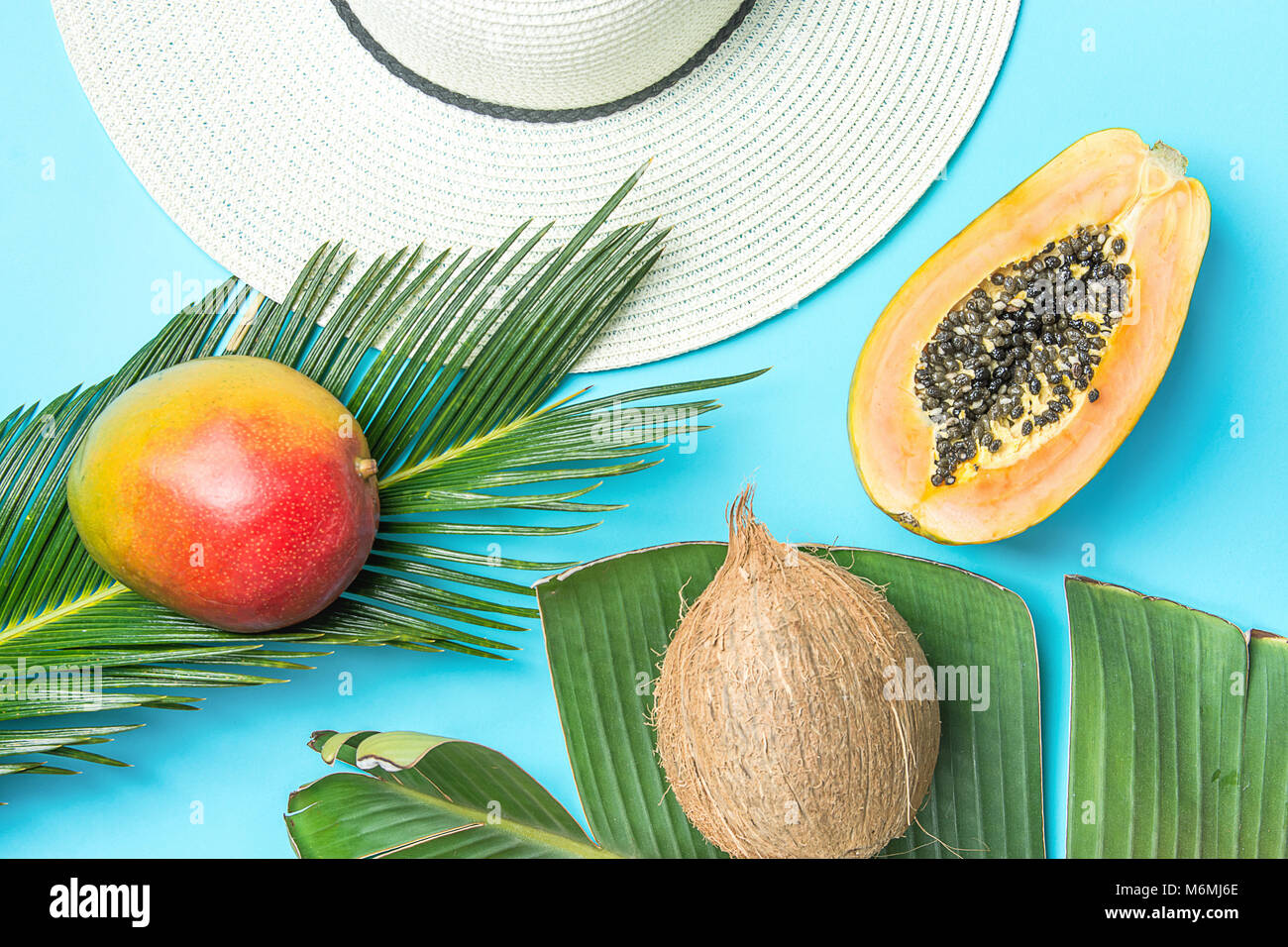 Ripe Juicy Mango Halved Papaya Coconut on Large Palm Leaf Straw Sun Hat on Blue Background. Summer Vacation Relaxation Fashion Concept. Healthy Lifest Stock Photo