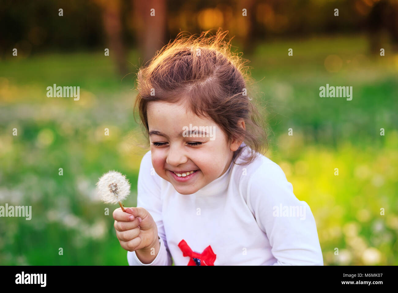 Beautiful little child with dandelion flower in sunny summer park. Happy cute kid having fun outdoors at sunset. Stock Photo