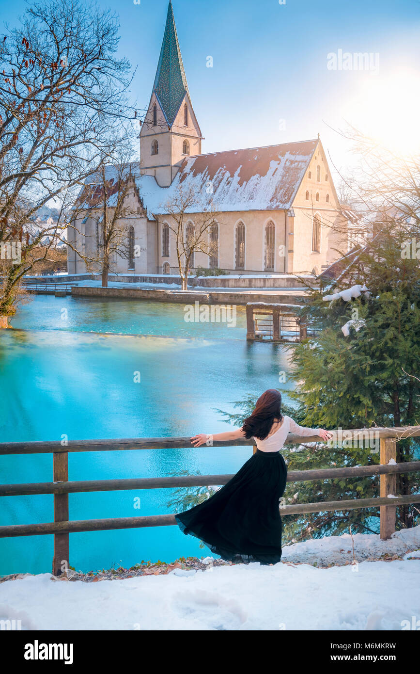 Rear view of an attractive young woman, dressed elegantly, admiring the blue water of the Blautopf spring, on a sunny day, in Blaubeuren, Germany. Stock Photo
