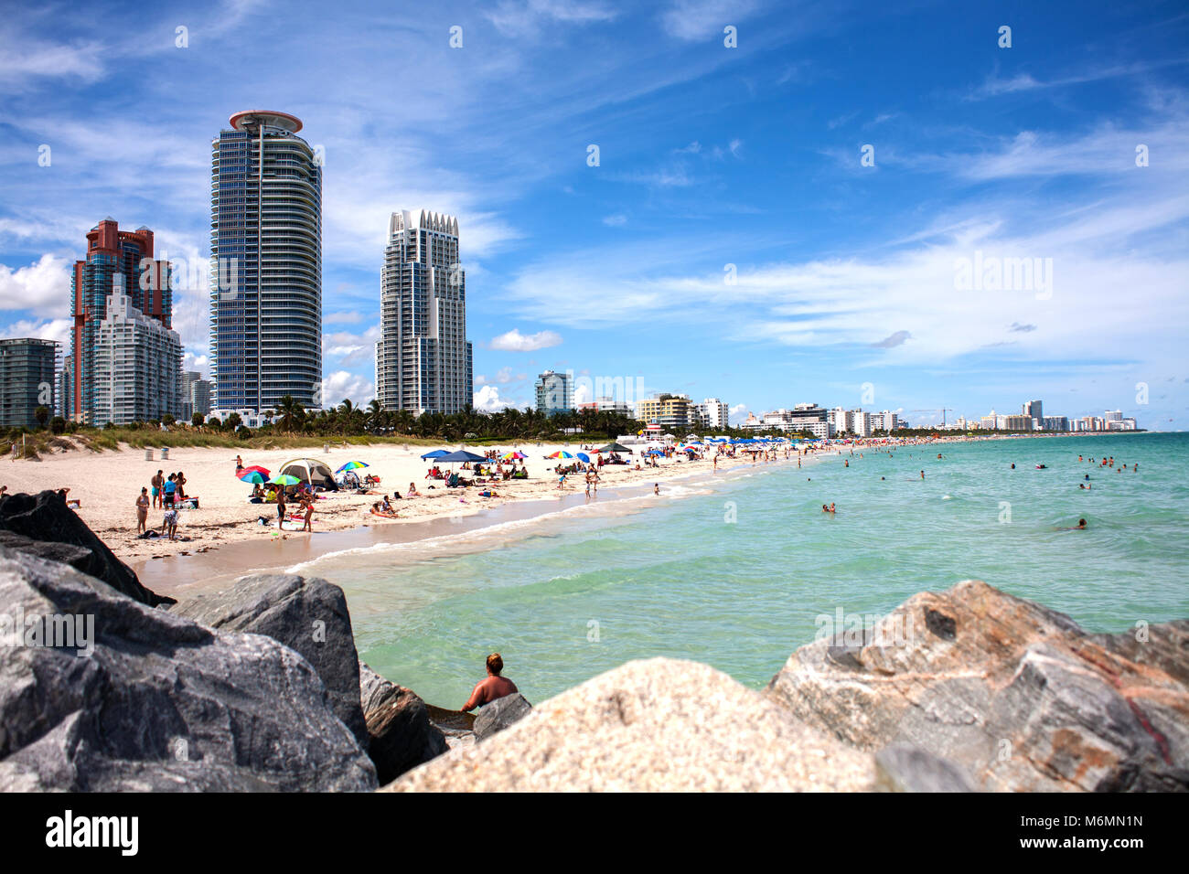 View of Miami South Beach over rocks during sunny day. Hotels and luxury apartments in background. People chilling on sandy beach. Stock Photo