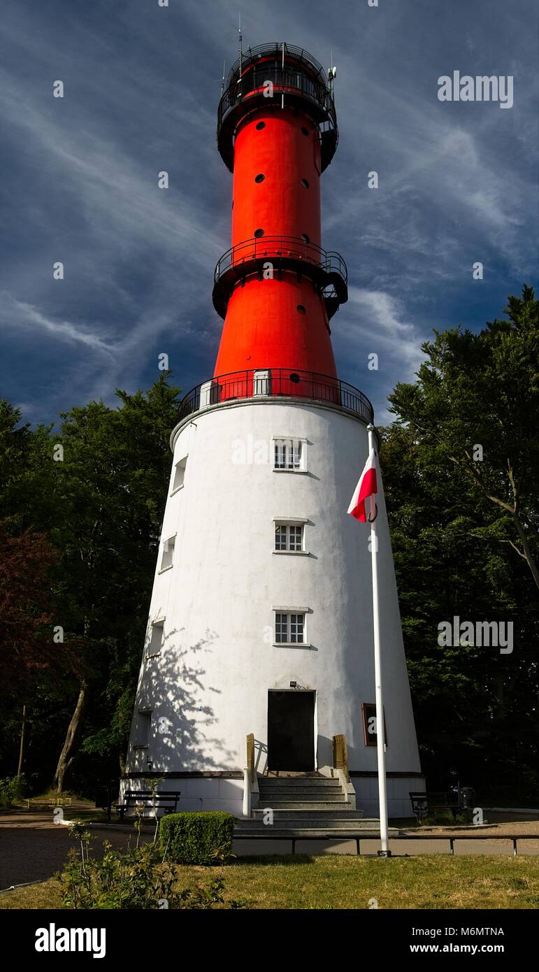 Red white lighthouse in sunny day. Surrounded by green trees / forest. Blue sky with clouds. Polish flag on post. Stock Photo