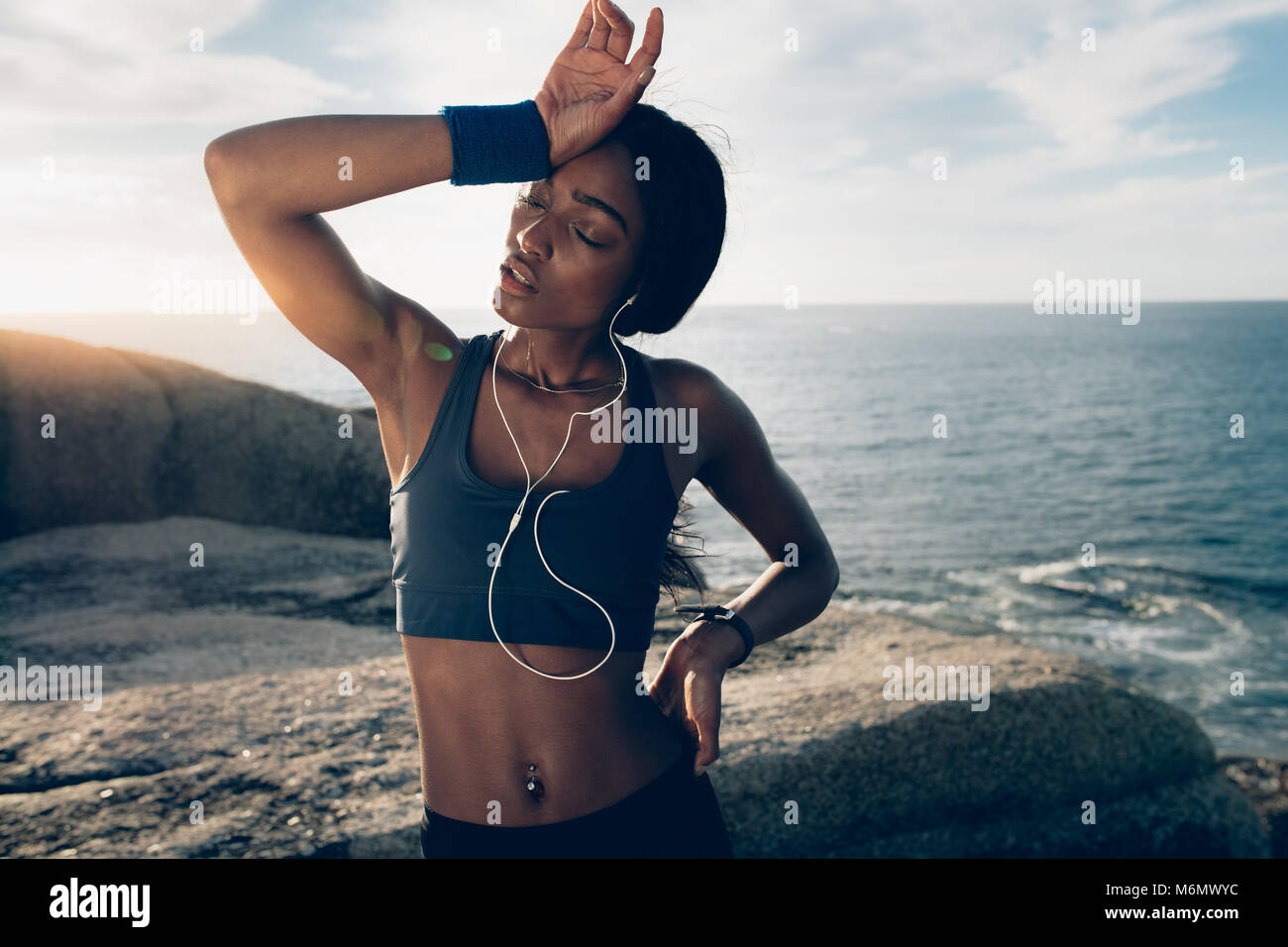 Fitness woman looking tired after intense physical training. Sportswoman standing over rocks at the beach with hand on head. Stock Photo