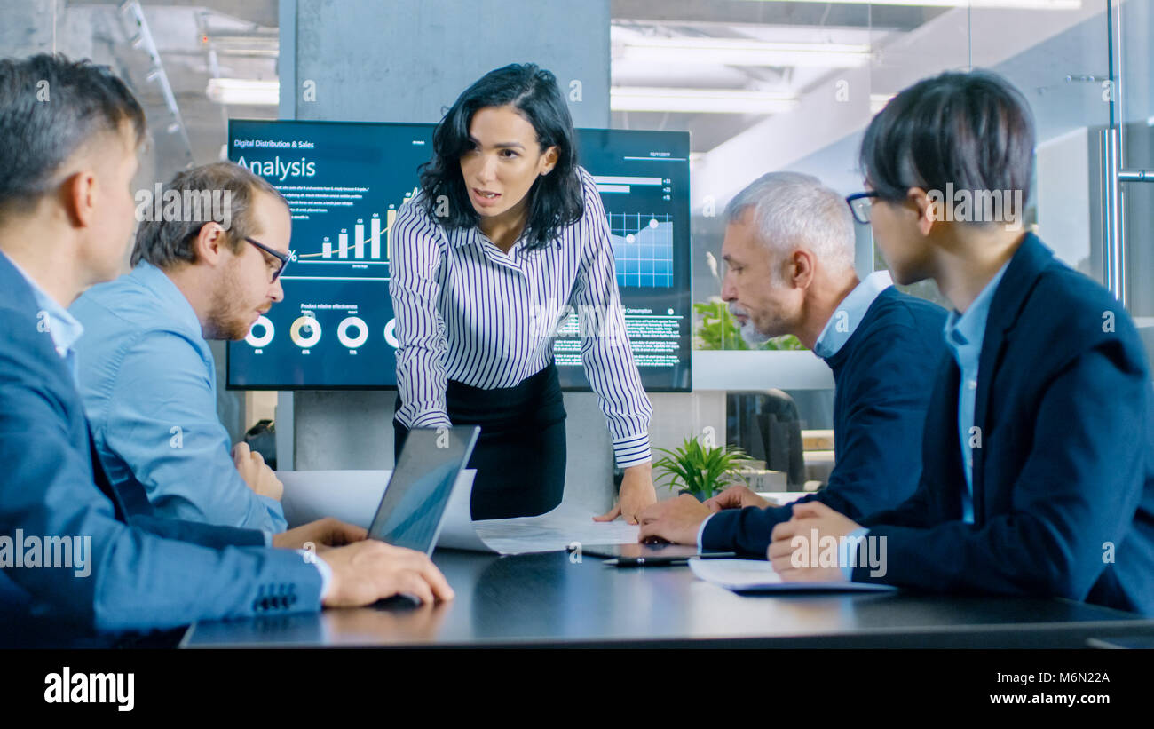 Chief Female Executive Leans and Spreads Project Blueprints on the Table Showing them to Her Colleagues. In the Background Pie Charts and Graphs Stock Photo