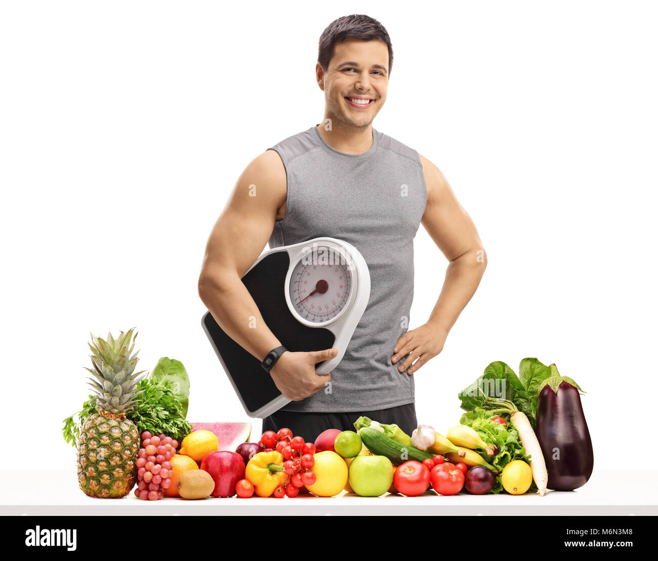 Young man dressed in sportswear holding a weight scale behind a table with fruit and vegetables isolated on white background Stock Photo