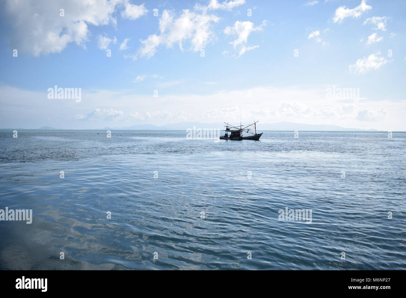 Small fisherman boat in the middle of the gulf of thailand alone with a blue sky and mountains in the background. Stock Photo