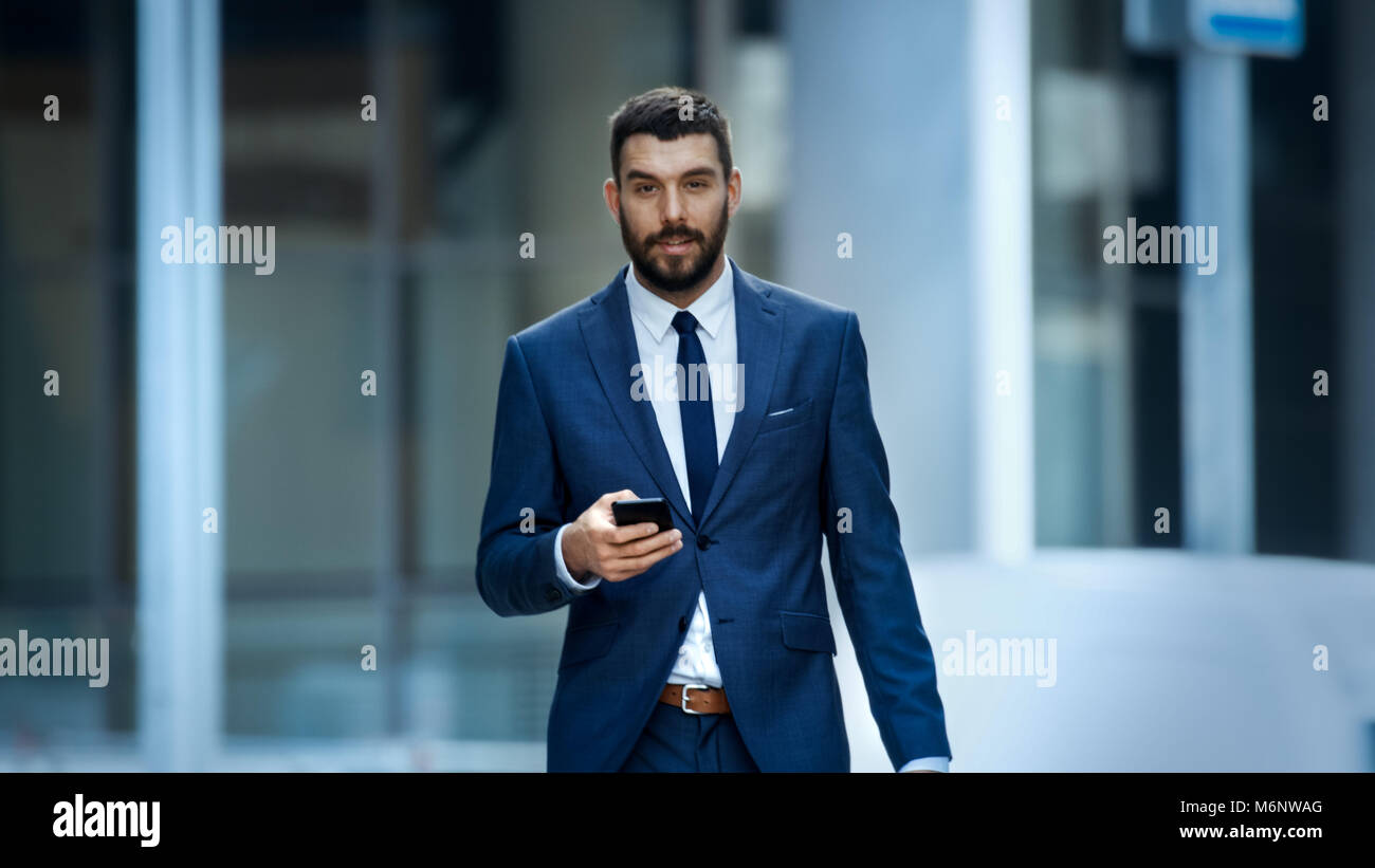Successful Business Man Uses Smartphone While Walking on the Big City Business District Street. He is looking into camera. Stock Photo