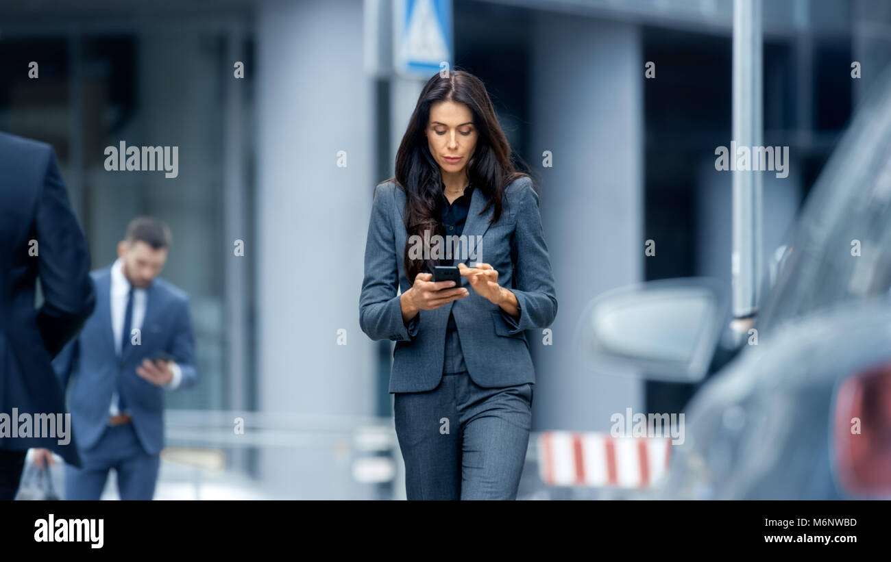 Business Woman in the Tailored Suit Walking on the Busy Big City Street in the Business District, Checks Her Smartphone. Confident Woman on Street. Stock Photo