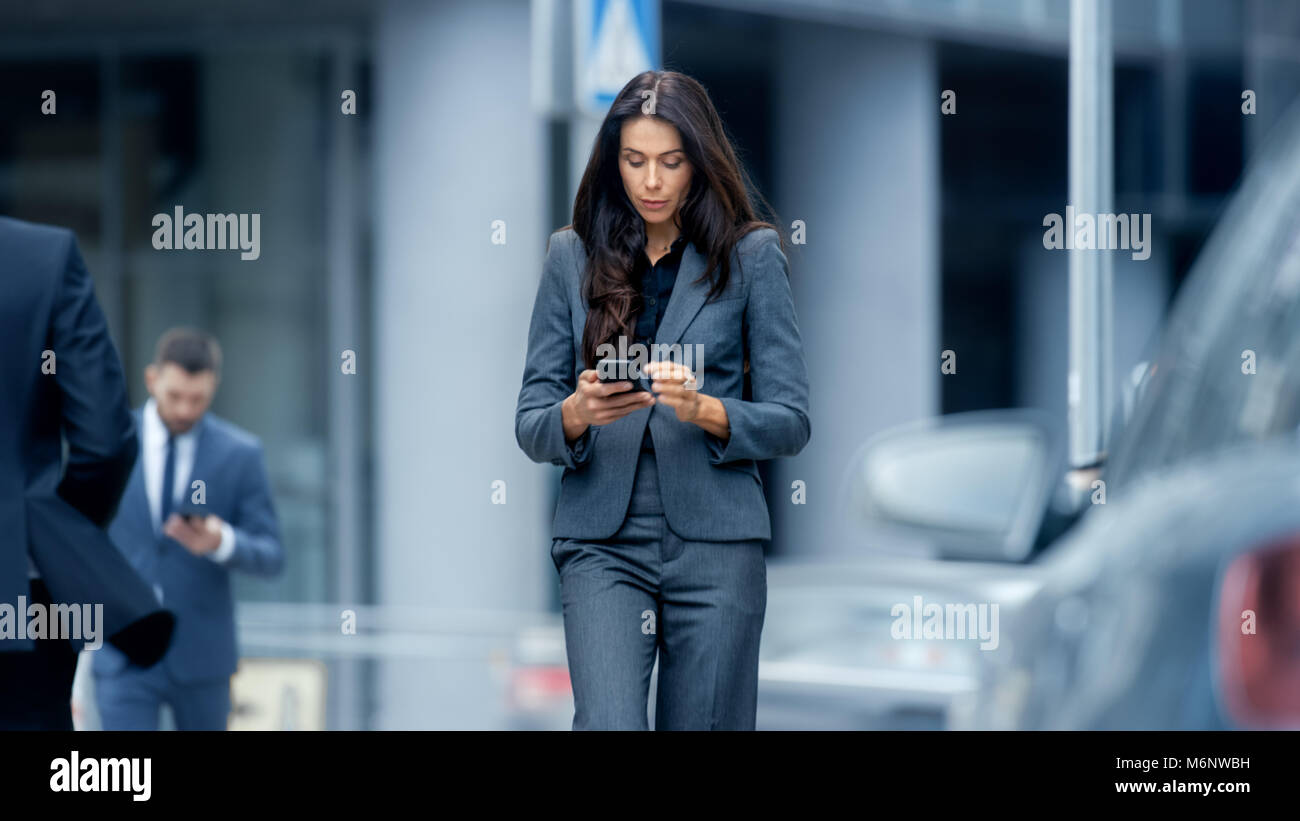 Business Woman in the Tailored Suit Walking on the Busy Big City Street in the Business District, Checks Her Smartphone. Confident Woman on Street. Stock Photo