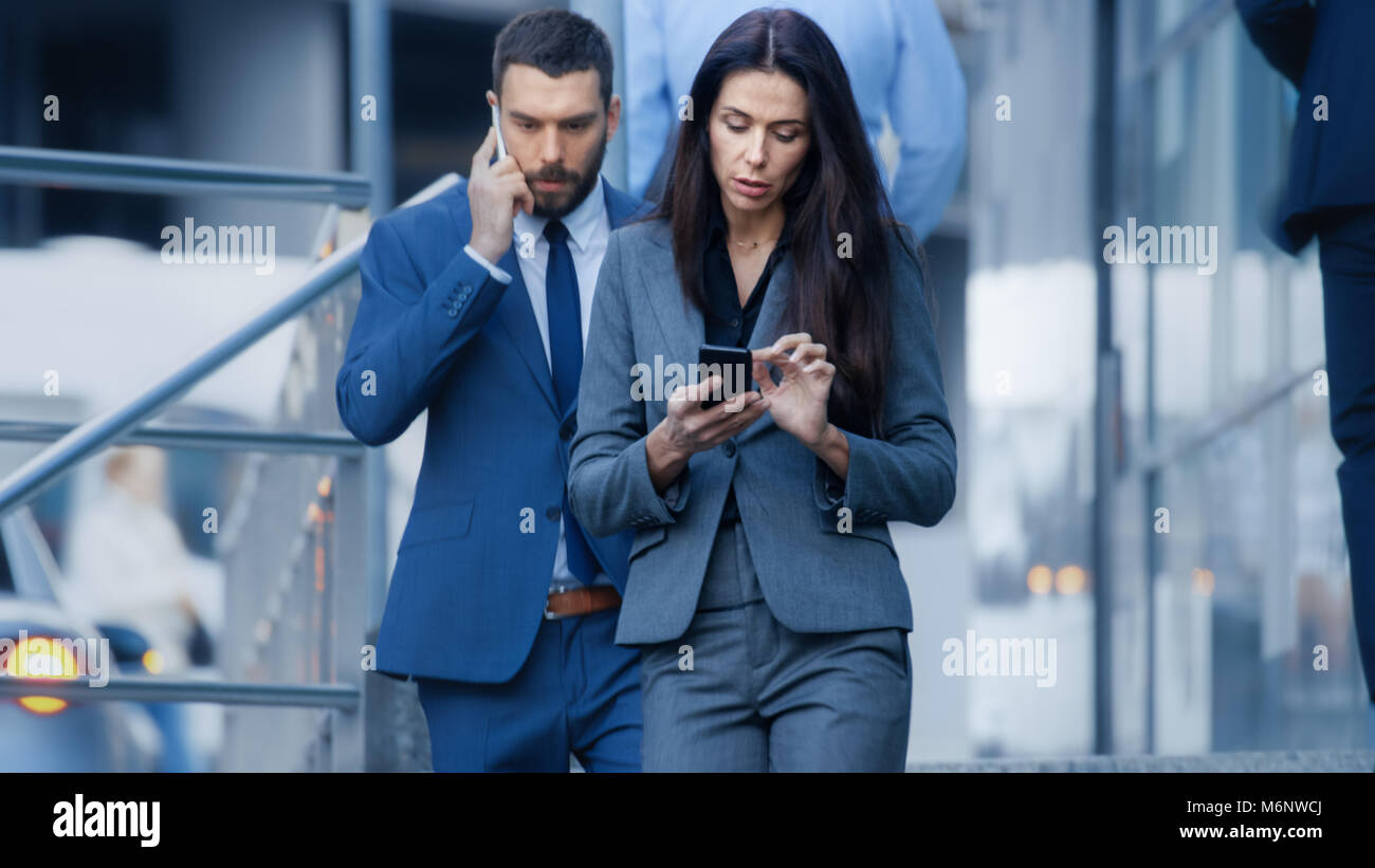 Male and Female Business People Walking on the Big City Business District Street. They both Wear Stylish Grey/ Blue Tailored Suits, Man Holds a Bag. Stock Photo