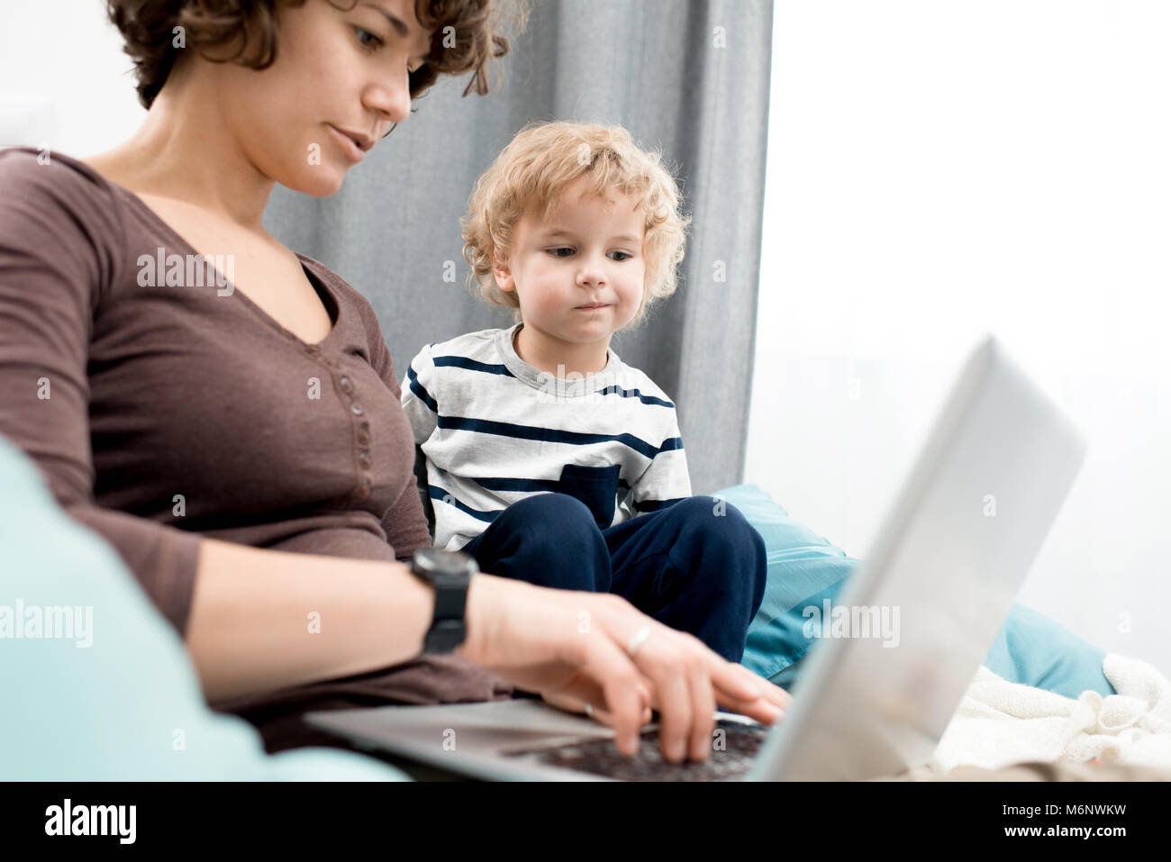 Portrait of young mother using laptop at home, focus on curious little boy looking at screen and learning about internet Stock Photo