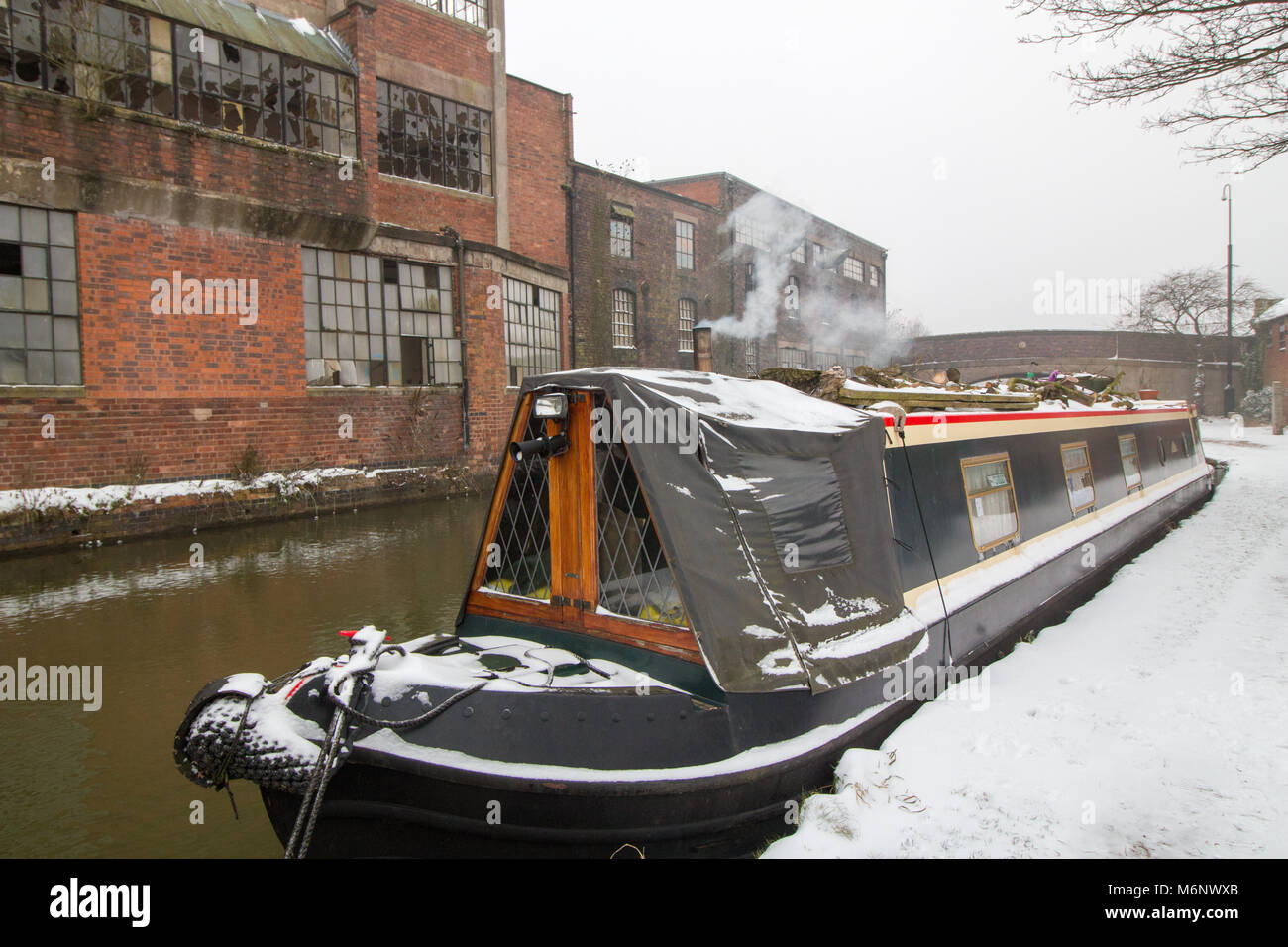 A canal boat covered in snow in front of the derelict  Britannia Works an old hat factory on the Coventry canal, near Atherstone, Warwickshire Stock Photo