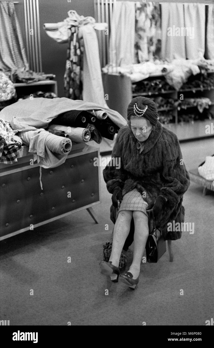 Fashionable middle aged older woman shopping, sitting down tired feet. She is wearing a real fur coat in the fabric department of a large London department store London UK 1971 1970s.  HOMER SYKES Stock Photo