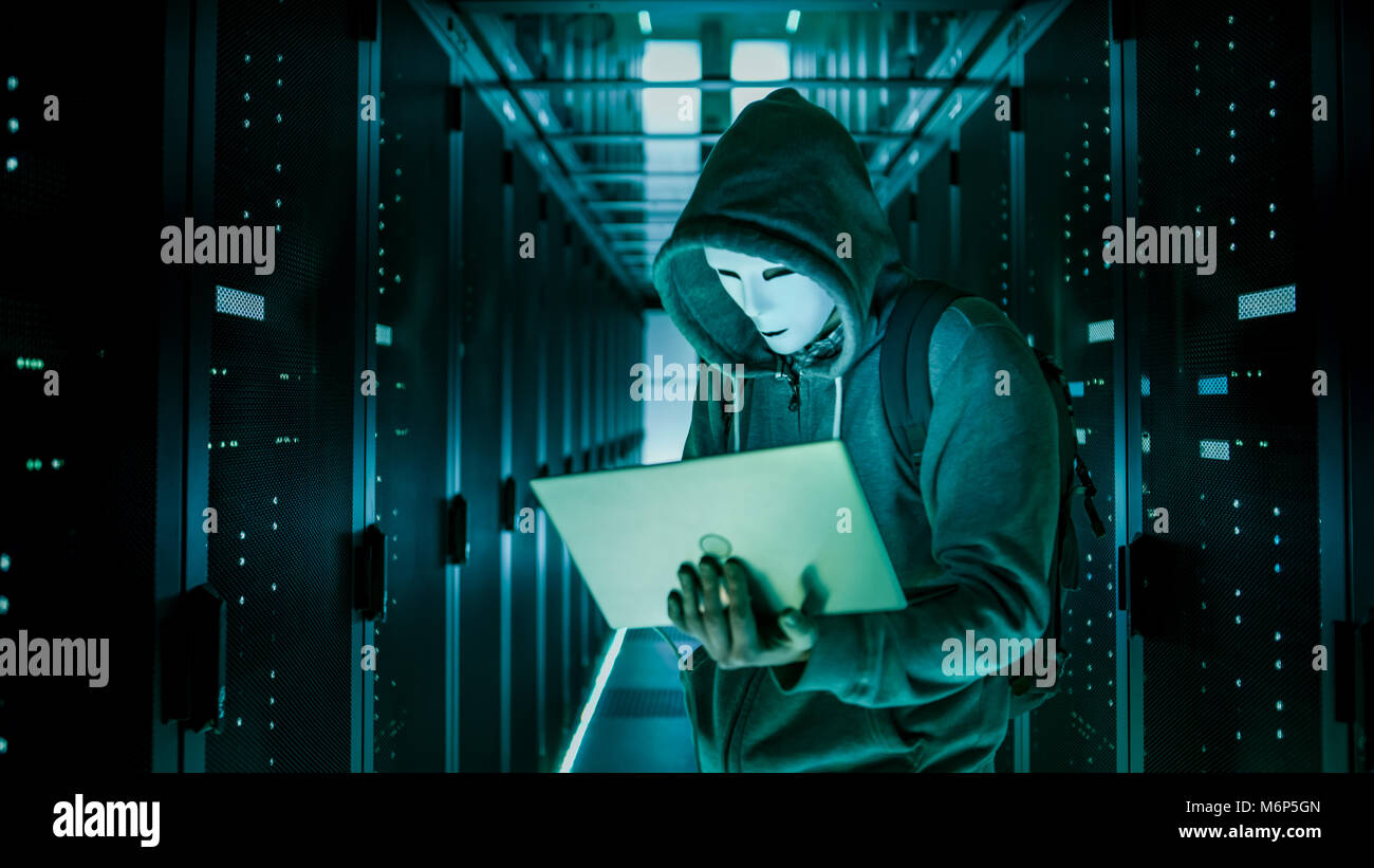 Mid Shot of a Masked Hacker in a Hoodie Standing in the Middle of Data Center full of Rack Servers and Hacking it with His Laptop. Stock Photo
