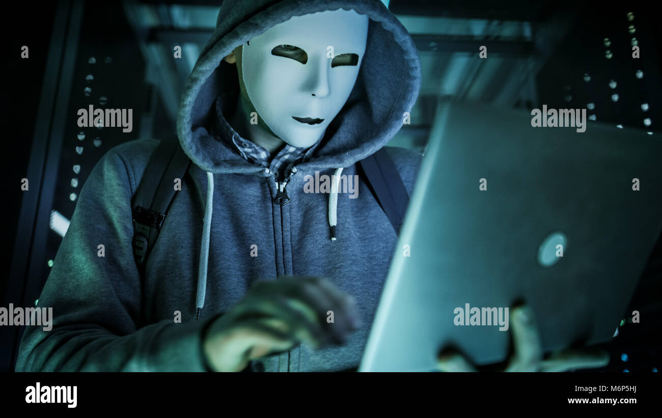 Close-up Shot of a Masked Hacker in a Hoodie Standing in the Middle of Data Center full of Rack Servers and Hacking it with His Laptop. Stock Photo
