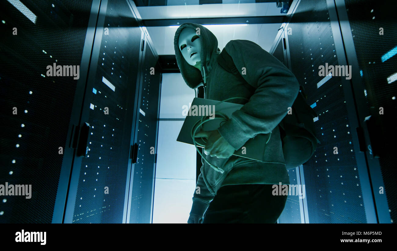 Low Angle Shot of a Masked Hacker in a Hoodie Sneaking Through Corporate Data Center with Rows of Rack Servers. Stock Photo