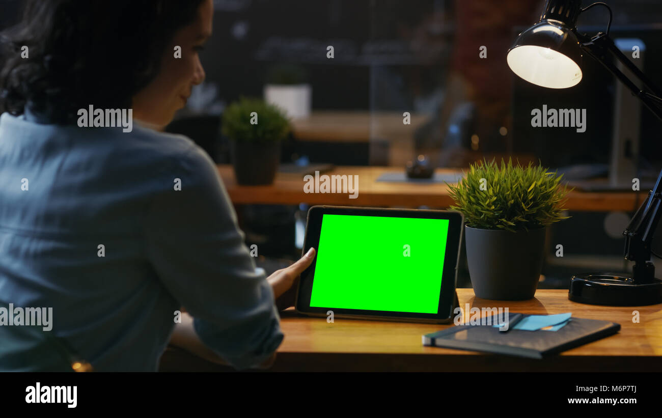 Young Woman Sits at Her Wooden Desk Uses Tablet Computer with Mock-up Green Screen, She Uses Various Swipe and Touch Gestures. Stock Photo
