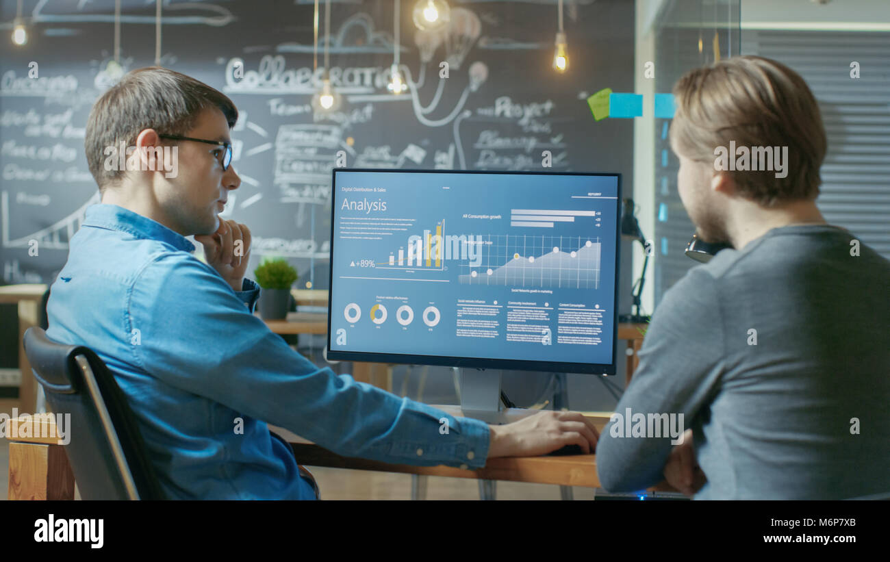 Two Office Employees Have Project Related Discussion, on the Desk Stands Personal Computer with Statistics, Graphs and Charts. Stock Photo