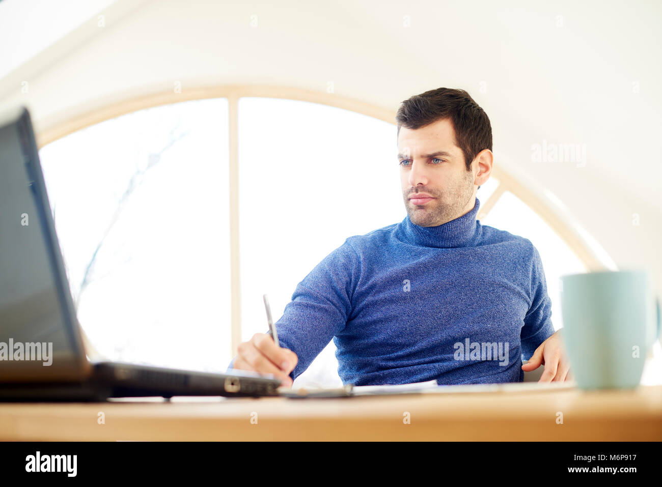 Portrait of casual young man wearing turtleneck sweater and looking thoughtul while working on laptop at home. Home office. Stock Photo