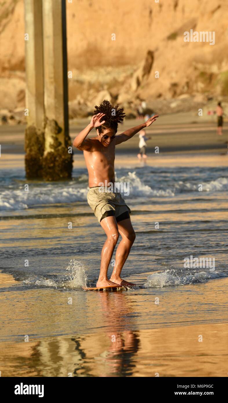 Young African-American man on a skim board, skimming, at beach during sunset, gliding across waves, San Diego, California, USA. Stock Photo