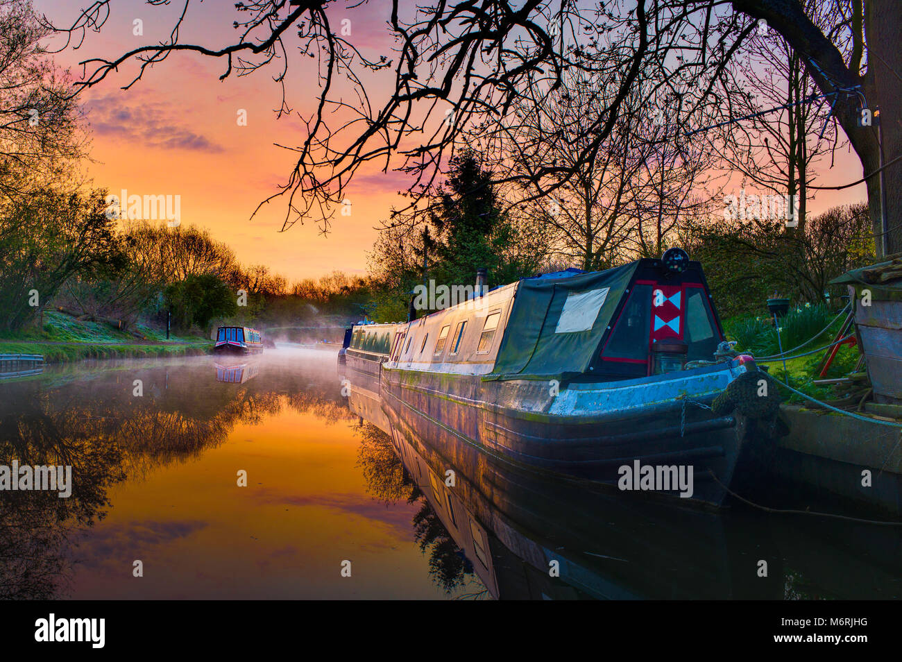 Tranquil reflections and morning mist in the first rays of dawn on the Grand union canal with several narrow boats. Stock Photo