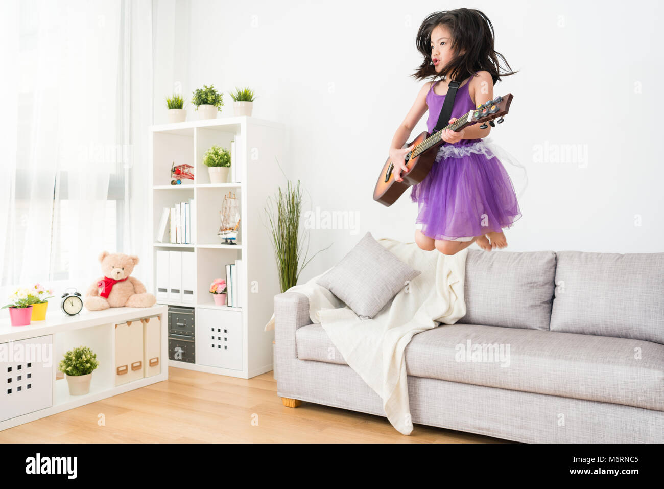 Little young girl daughter jumping crazily on the sofa with a wooden guitar. Thinking she is the real rocker. Stock Photo