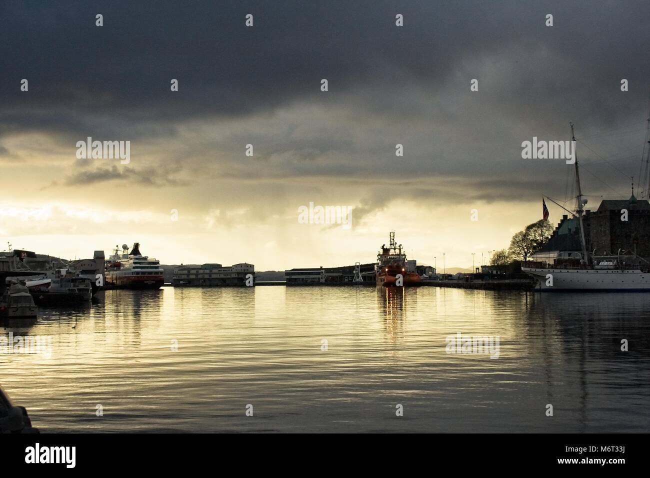 Bergen port. Dramatic look at the water ships and ferry. Rainy clouds. Sun reflection in the water. Stock Photo