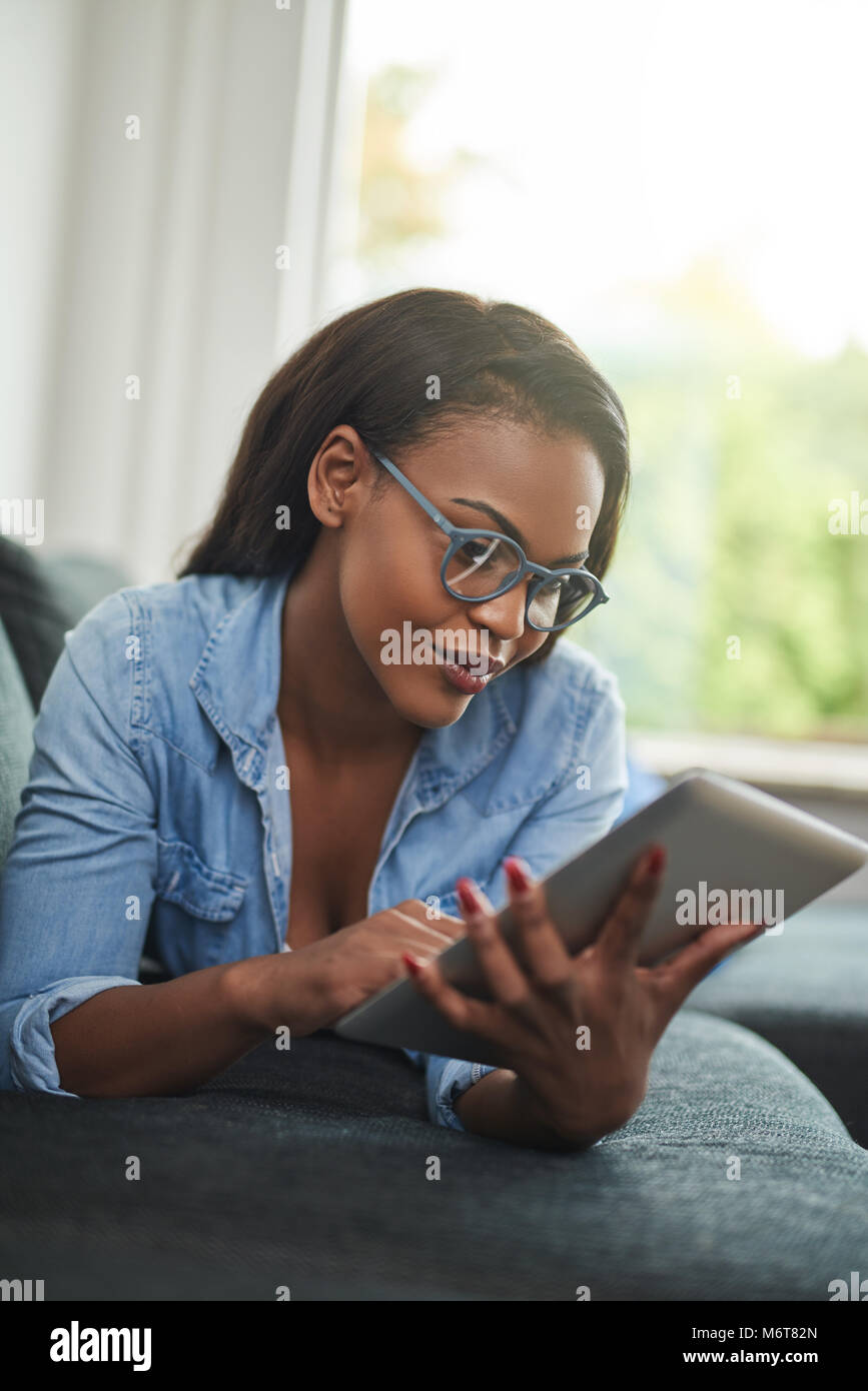 Smiling young African woman lying down on her living room sofa using a ...