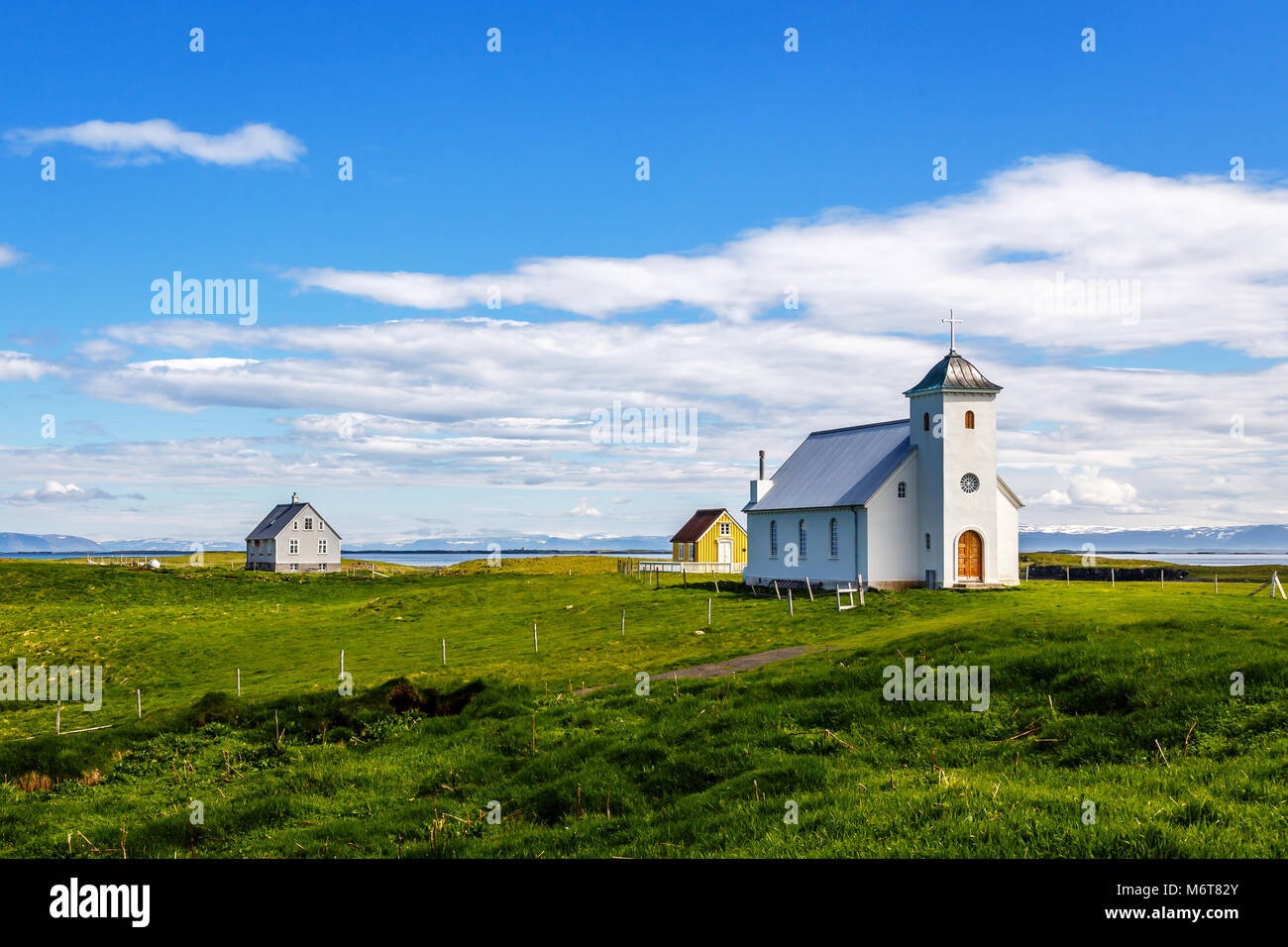 Flateyjarkirkja church and couple of living houses with meadow in foreground and sea  fjord with blue sky in the background, Flatey, Iceland Stock Photo