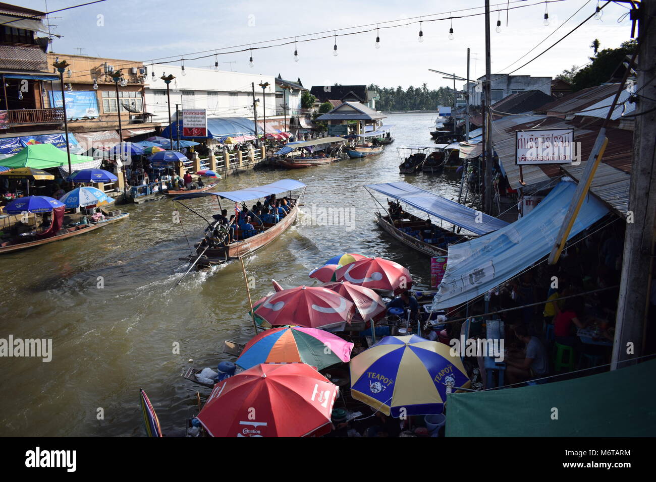 Wide view from the main bridge of a wooden tour boat  crossing the Amphawa floating market with many colorful umbrellas over merchants cooking food. Stock Photo