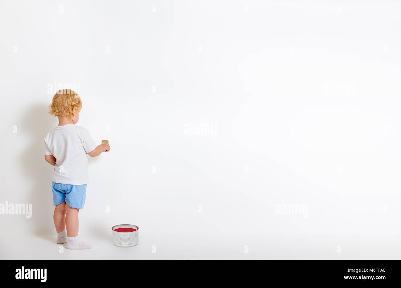Little boy with paint brush and tin can standing back near white wall Stock Photo