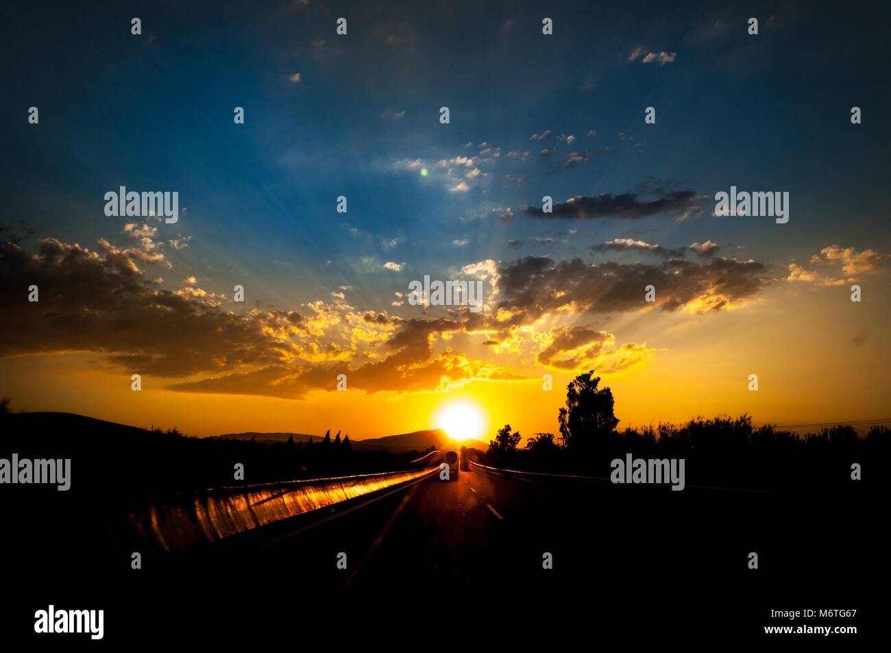Colorful sunset over at highway. Landscape with orange sunset over highway. Cloud layers with sunrays between them. Contre-jour silhouette of cars. Stock Photo
