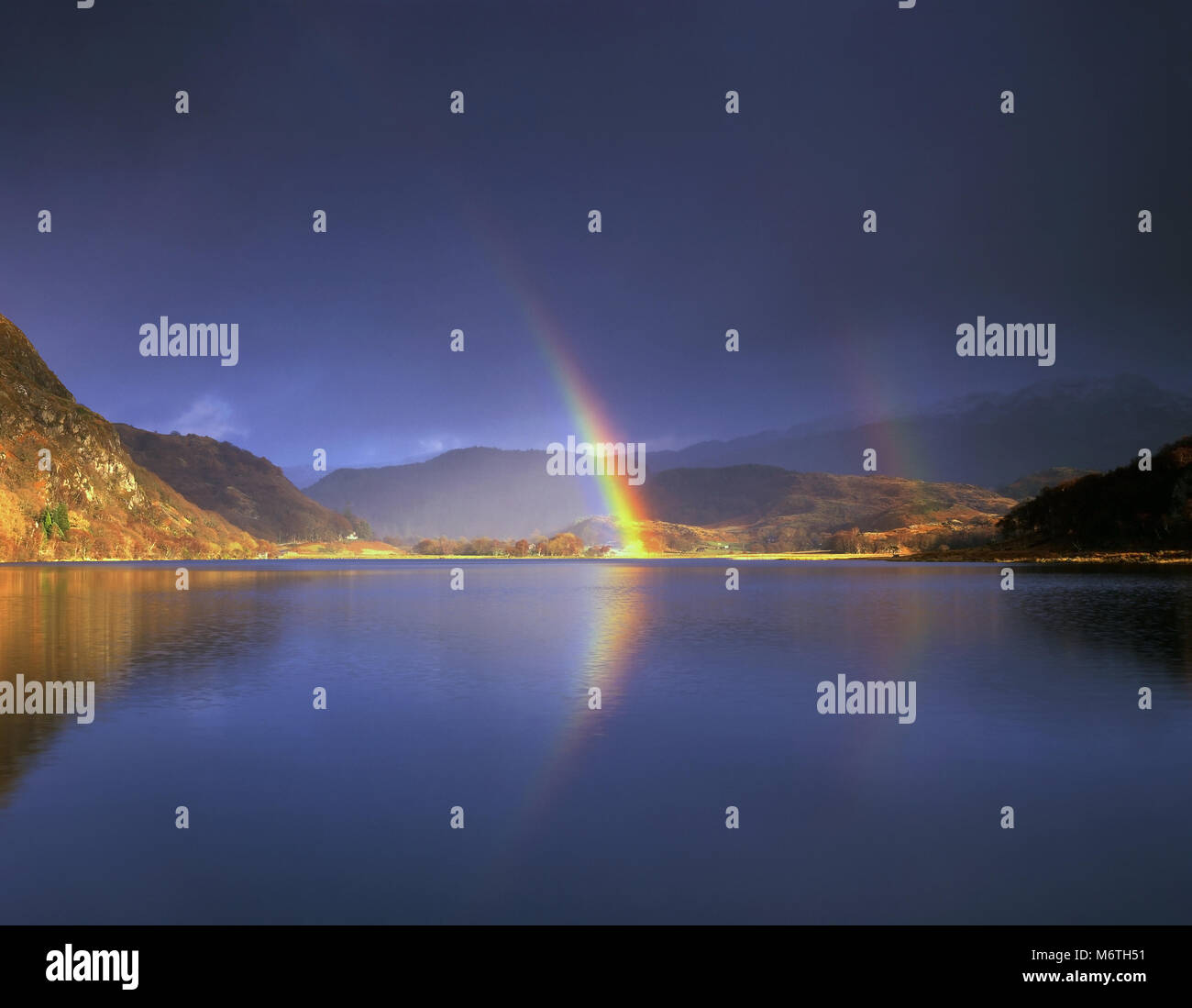 A vivid rainbow brightens up a dark sky over Llyn Dinas in the Snowdonia National Park, Wales. Stock Photo
