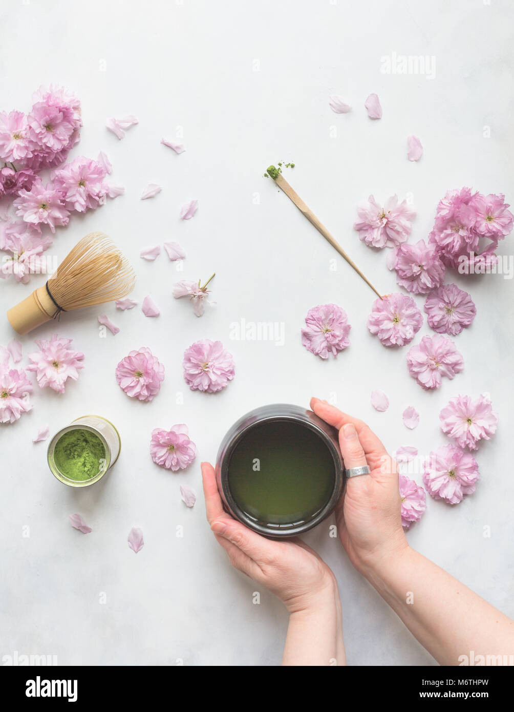 view from above of two female hands holding a cup of green Matcha tea, with cherry flowers, whisk and measuring spoon. Stock Photo