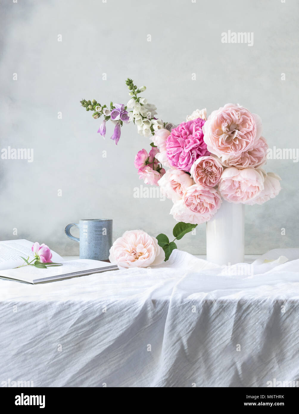 Still life, indoor, front view of table covered with pale grey linen, with white vase of pink David Austin roses & foxgloves, pale blue mug, open book Stock Photo
