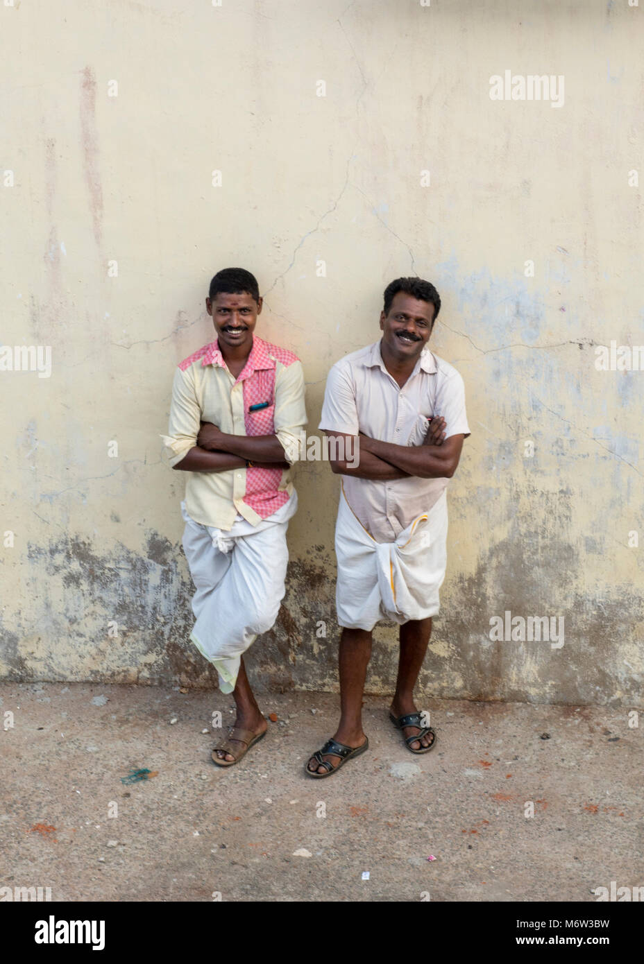 Two smiling men leaning against a wall smiling in Vizhinjam Fishing Village, Kerala, India Stock Photo