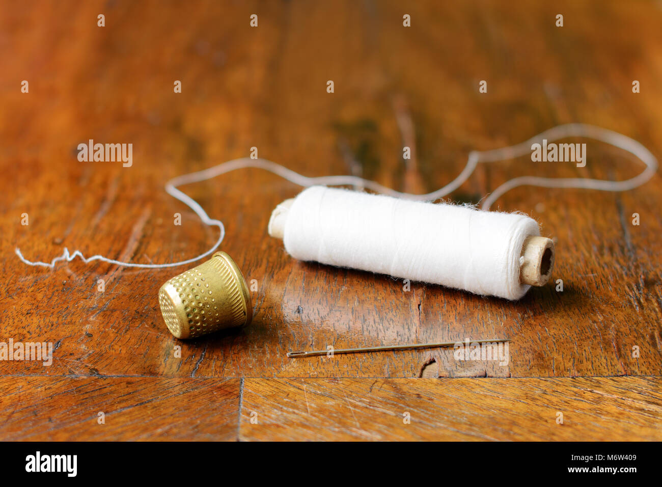 Thimble, a needle and a spool of white thread on an antique wood veneer full of cracks. Selective focus. Closeup. Stock Photo