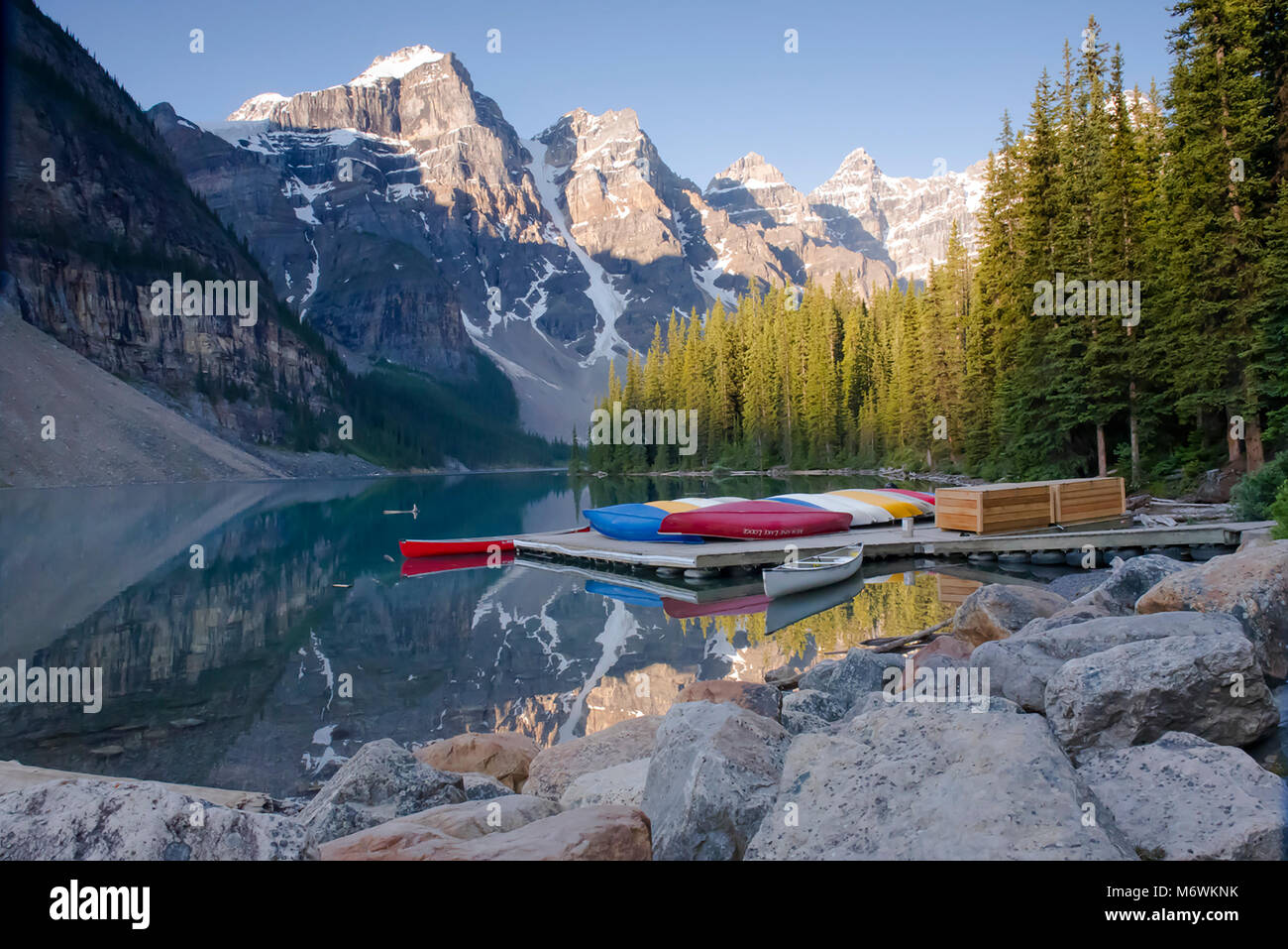 Brightly coloured canoes on a jetty, Lake Moraine, Alberta, Canada. The morning light illuminates the snow capped peaks Stock Photo