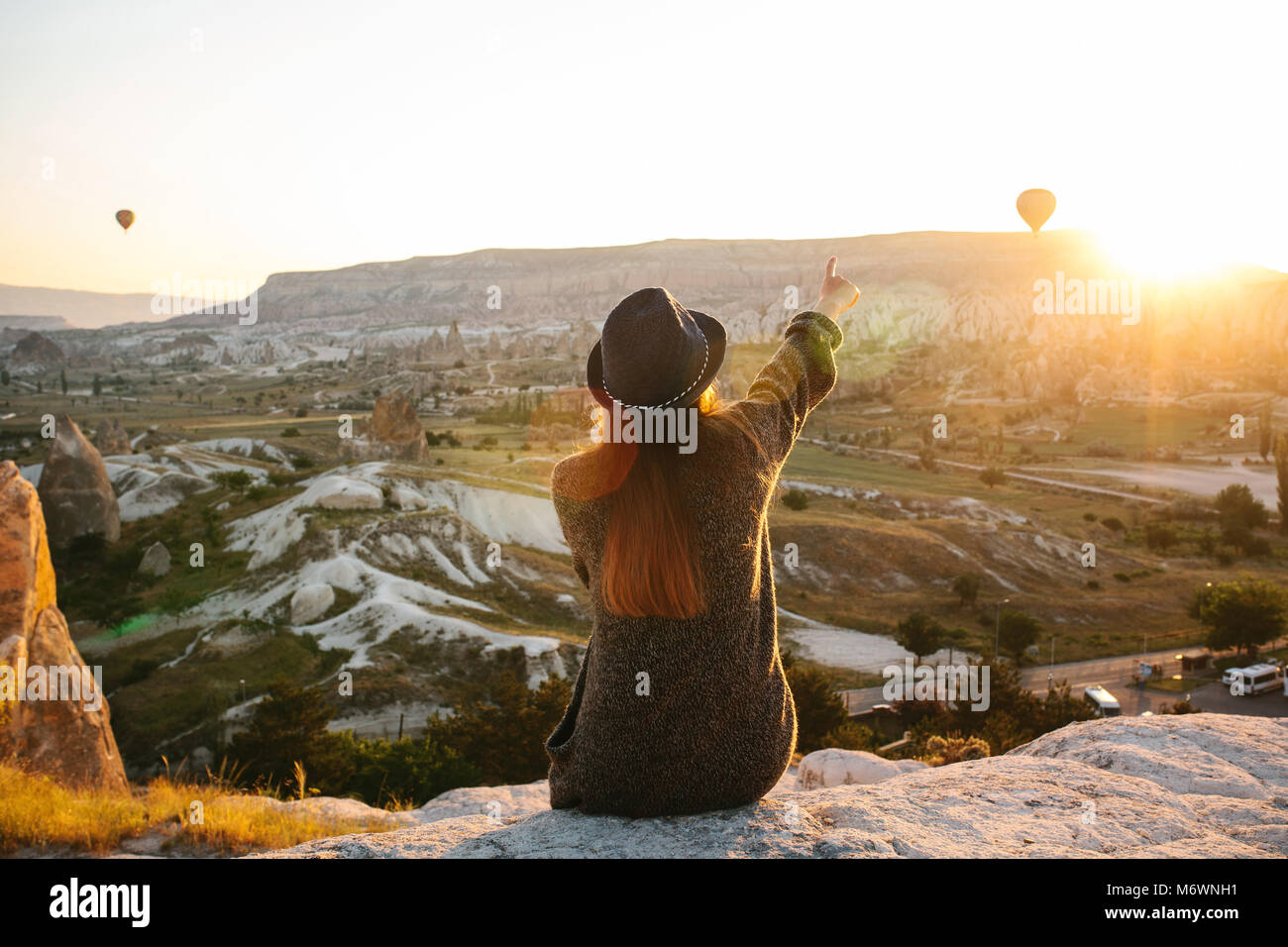 A tourist girl in a hat sits on a mountain and looks at the sunrise and ...