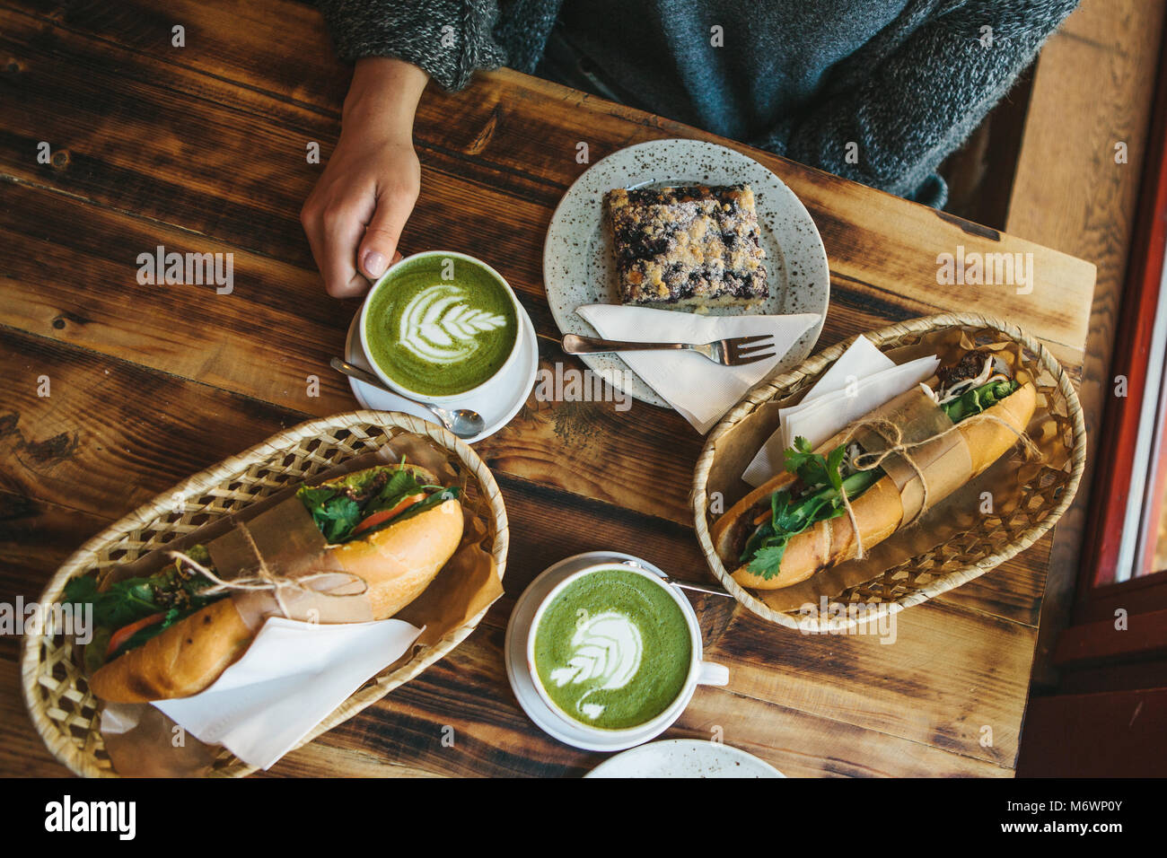 Top view - The girl sitting in cafe and holding mug with green tea with milk next to piece of sweet pie and two sandwiches with vegetables. Stock Photo