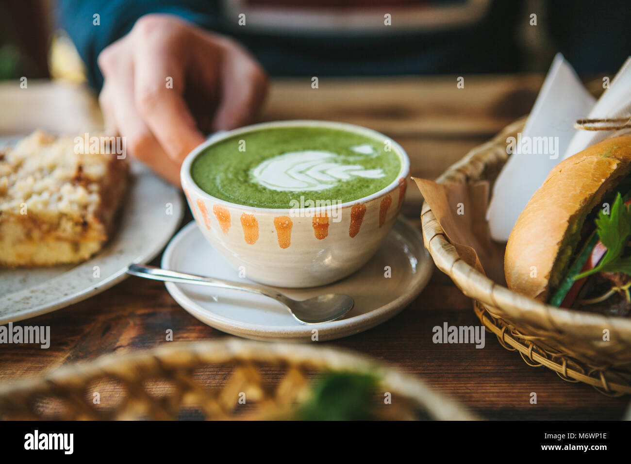 Close-up man's hand holding mug of green tea with beautiful pattern in the form of white foam next to dessert and sandwich Stock Photo