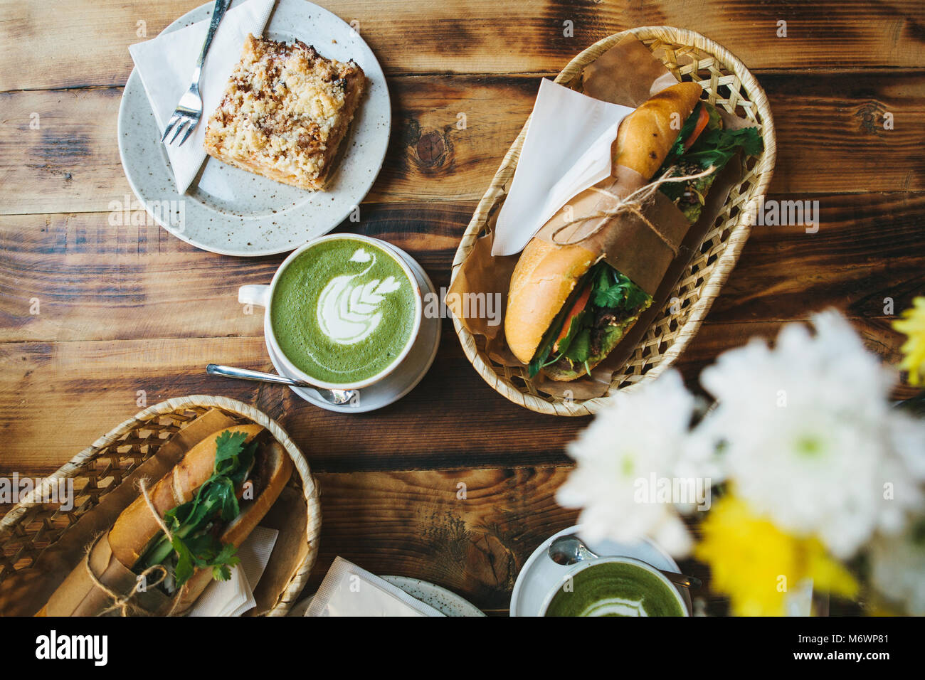Healthy breakfast in the restaurant: cups of green tea with milk called Matcha, dessert and sandwiches with vegetables and herbs on wooden table. Nearby are yellow flowers to decorate the table. Stock Photo