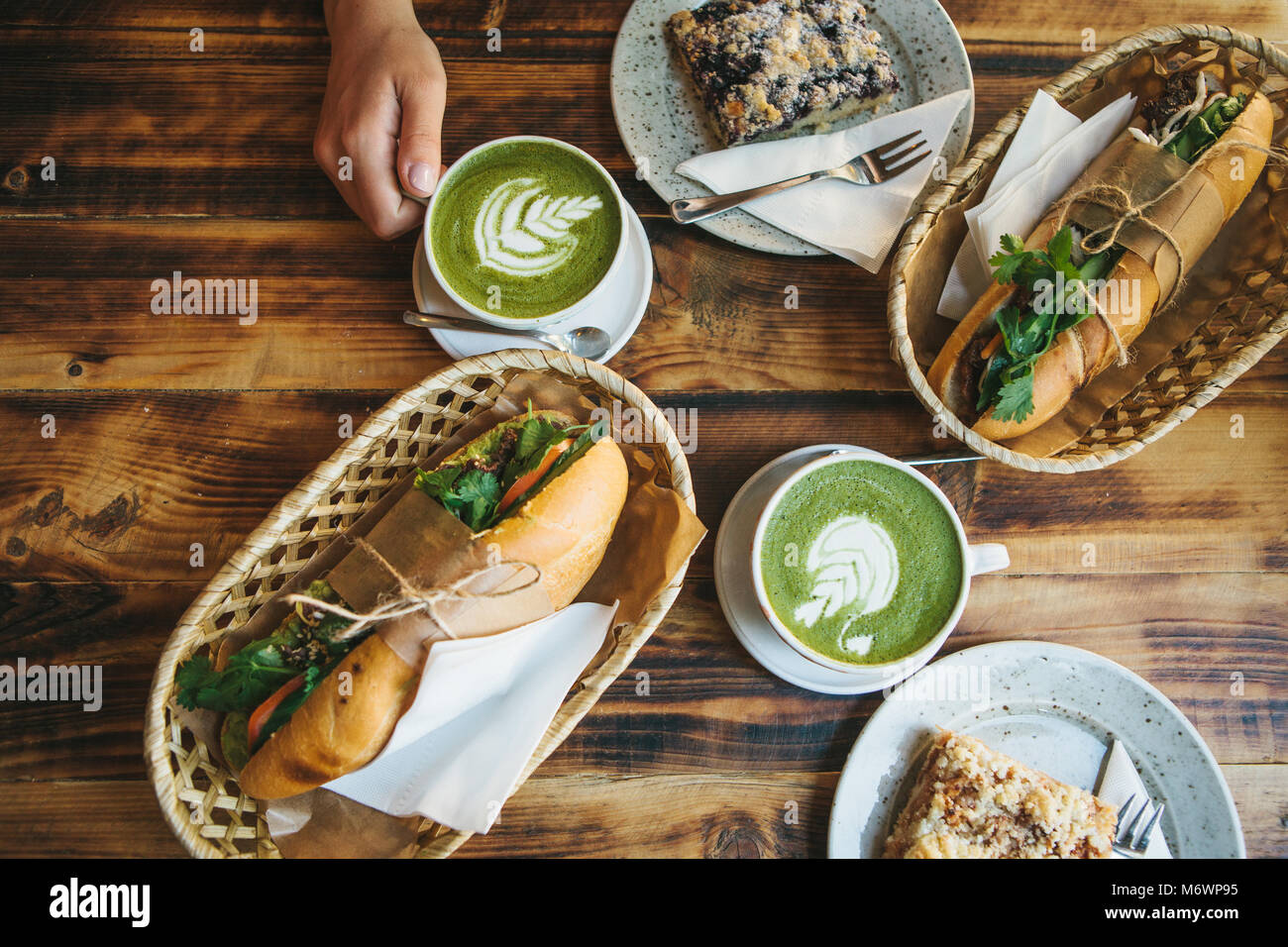 Top view - The girl sitting in cafe and holding mug with green tea with milk next to piece of sweet pie and two sandwiches with vegetables. Stock Photo