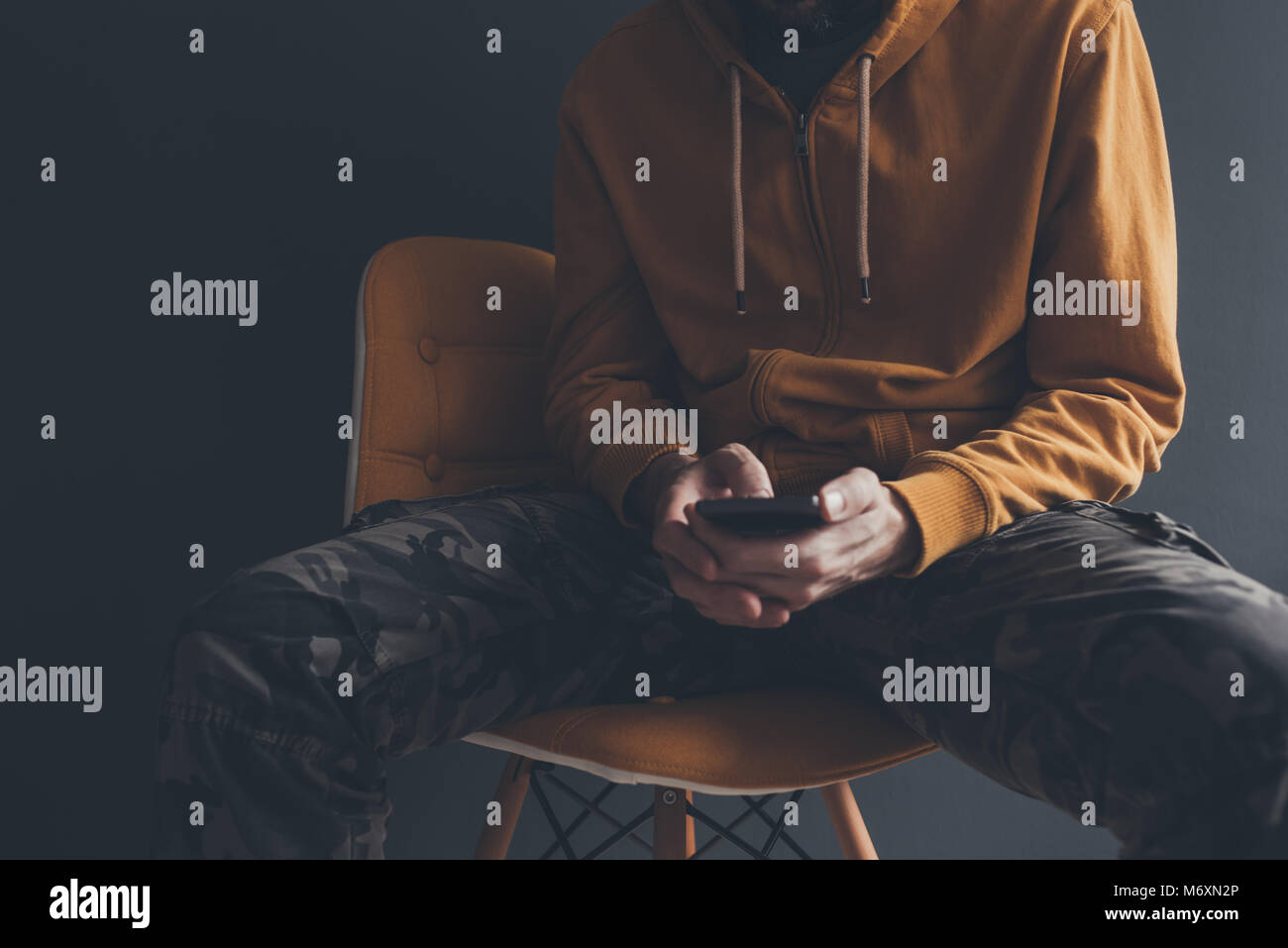Casual man using smart phone laid back in office chair. Male person wearing yellow hoodie and military trousers is playing with his mobile phone. Stock Photo