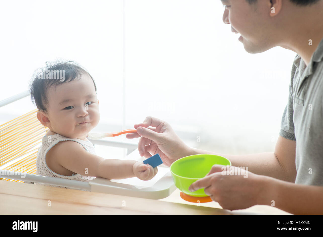 Happy Asian family at home. Father feeding solid food to his 9 months old child in the kitchen, living lifestyle indoors. Stock Photo