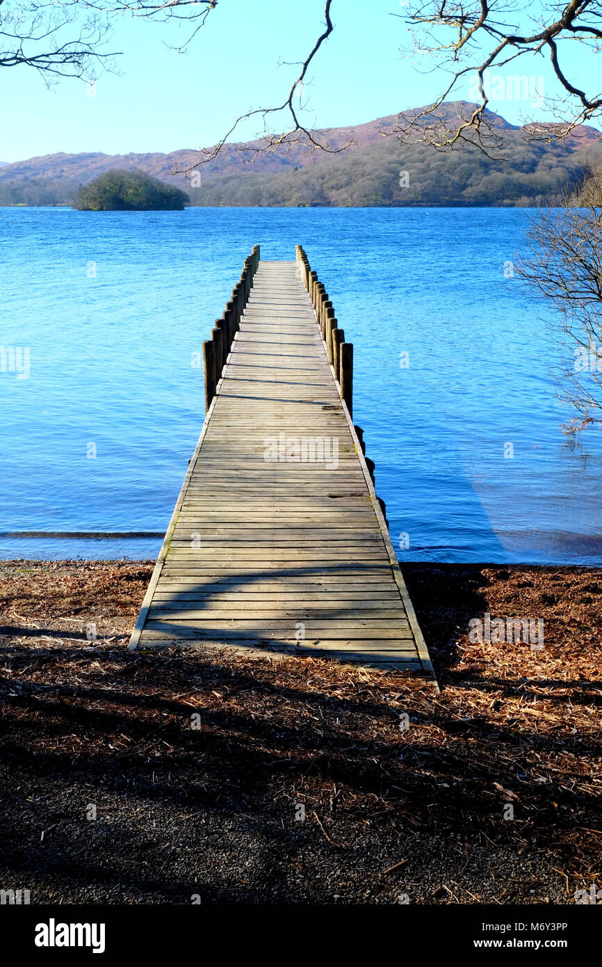 Long straight wooden foot jetty jutting out over a calm blue lake, with hills of green fields and forest woodland in the background, the sun is shinin Stock Photo