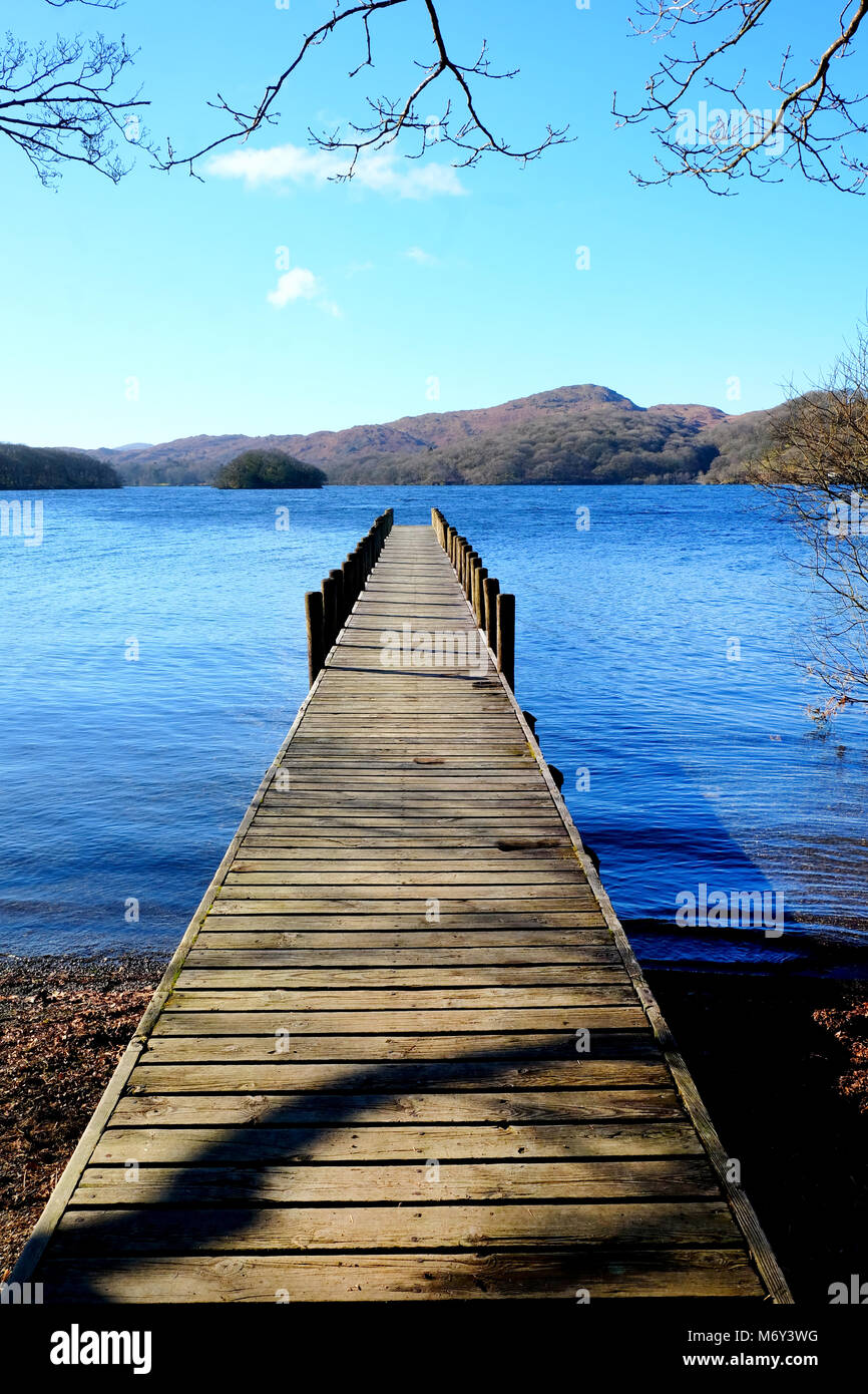 Long straight symetrical wooden foot jetty jutting out over a calm blue lake, with hills of green fields and forest woodland in the background, the su Stock Photo