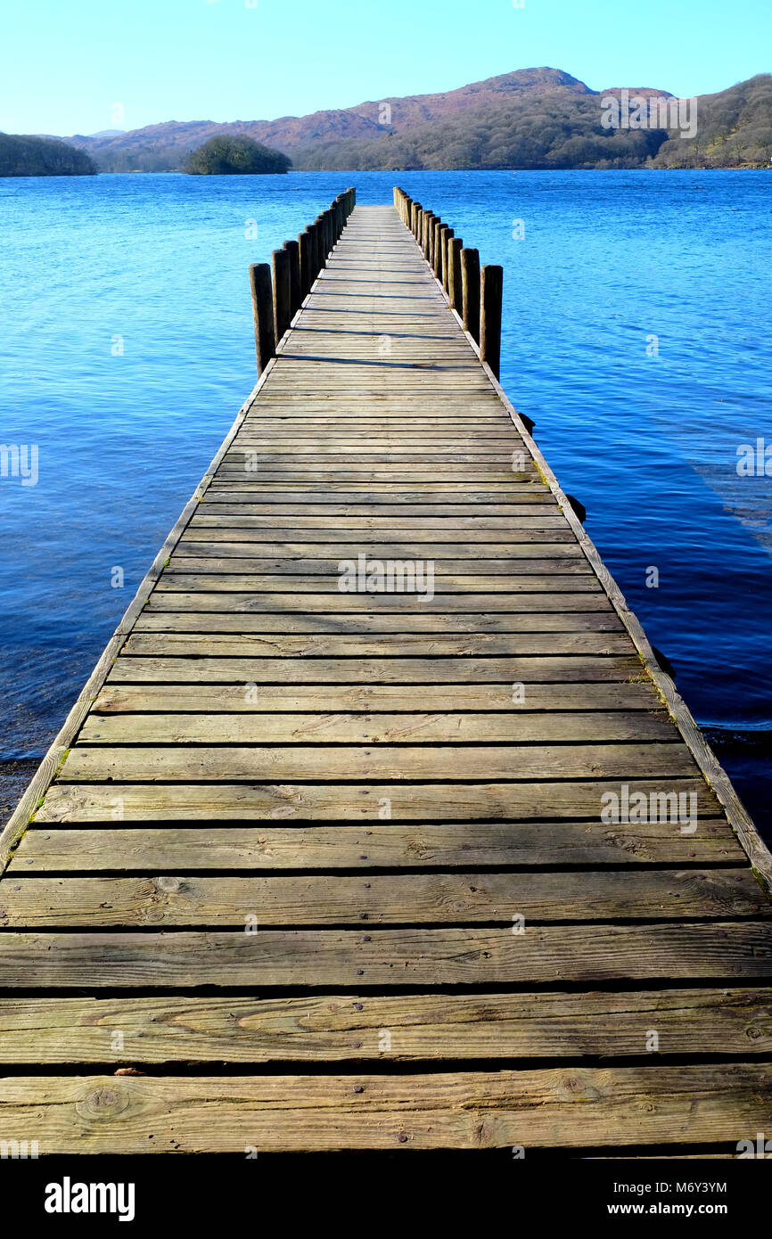 Very Long straight symetrical wooden foot jetty jutting out over a calm blue lake, with hills of green fields and forest woodland in the background, t Stock Photo