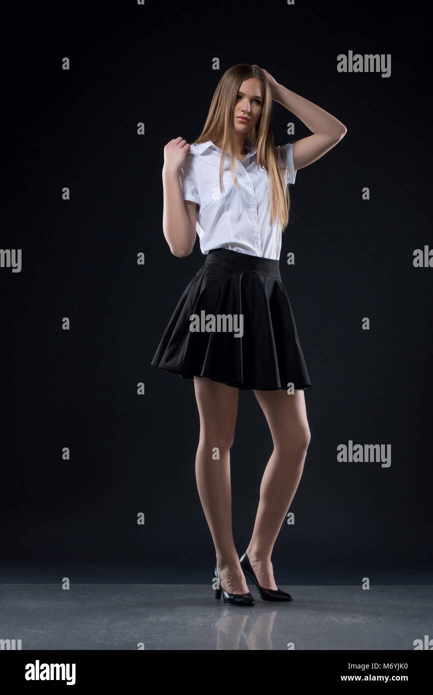 studio portrait of beautiful blonde caucasian young woman teen girl on black background posing in a white blouse and short black skirt, office look. H Stock Photo